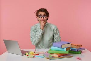 Positive young male with wild hair sitting at working table with books and laptop, posing over pink background in shirt and glasses, holding chin with hand and looking to camera with raised eyebrows photo