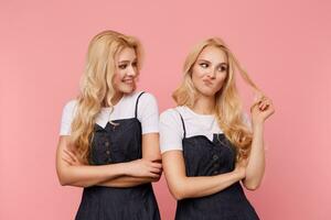 Glad young attractive female with wavy long hair looking cheerfully on her puzzled pretty sister grimacing face and pulling hair with raised hand, isolated over pink background photo