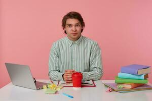 Portrait of handsome young dark haired man in striped shirt sitting at table with folded arms, looking to camera with calm face, posing over pink background photo