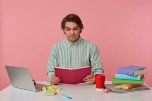 Indoor photo of concentrated young dark haired man sitting at working table with book in hands, biting underlip and raising eyebrows, isolated over pink background