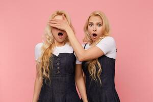 Studio shot of frightened long haired blonde women dressed in jeans dresses keeping their mouths wide opened while standing against pink background photo