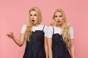 Horizontal shot of young bewildered long haired white-headed females keeping their mouths opened while looking confusedly aside, isolated over pink background photo