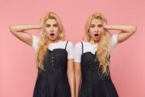 Shocked young lovely long haired blonde women keeping raised palms on their heads while looking dazedly at camera with wide mouths opened, isolated over pink background photo