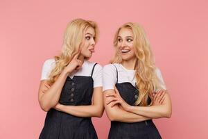 Two beautiful young white-headed women dressed in jeans dresses and white t-shirts being in nice mood and having fun together while posing over pink background photo