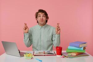 Portrait of cute young dark haired male raising hands with crossed fingers, making a wish to pass exams, posing over pink background in striped shirt photo