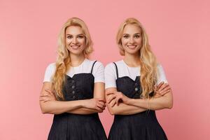 Happy young beautiful white-headed ladies folding hands on their chests while looking positively at camera with charming smiles, isolated over pink background photo