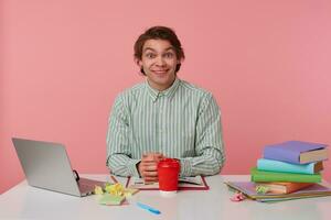 estudio Disparo de alegre linda joven masculino haciendo descanso con estudiando y Bebiendo café, sonriente extensamente a cámara con elevado Cejas, posando terminado rosado antecedentes foto