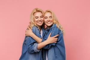 Good looking young attractive long haired blonde ladies keeping eyes closed while embracing each other lovingly, isolated over pink background in casual wear photo