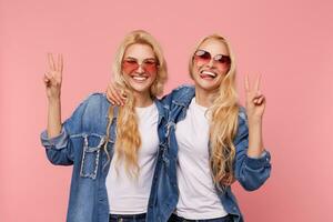 Happy young beautiful long haired blonde ladies in sunglasses and casual wear raising hands with victory signs and looking cheerfully at camera, isolated over pink background photo