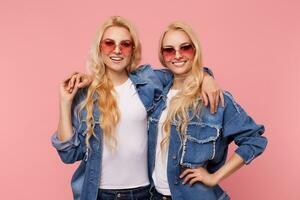 Charming young cheerful long haired white-headed sisters in red sunglasses hugging lovingly each other and looking happily at camera, standing over pink background photo