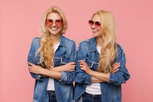 Happy young long haired blonde females dressed in sunglasses and casual clothes keeping hands folded and laughing cheerfully while posing over pink background photo