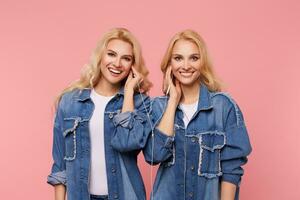 Happy young attractive long haired blonde sisters looking at camera with charming smiles while listening to music in headphones, isolated over pink background photo