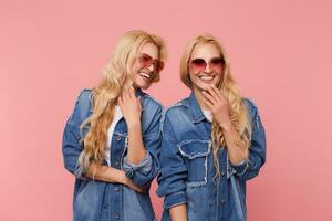 Indoor shot of young lovely happy long haired blonde twins dressed in same clothes having nice time together and laughing happily while standing over pink background photo
