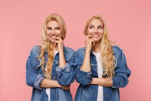 Indoor photo of young long haired blonde women looking gladly aside with wide smile and keeping forefingers on their lips, isolated over pink background