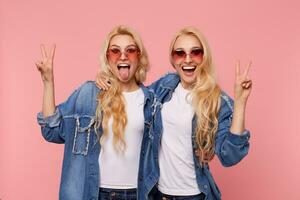 Studio shot of young joyful pretty long haired sisters dressed in jeans coats showing peace signs and looking joyfully at camera while posing over pink background photo