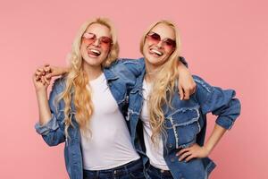 Happy young lovely long haired blonde ladies dressed in casual clothes laughing happily while embracing each other, standing against pink background photo