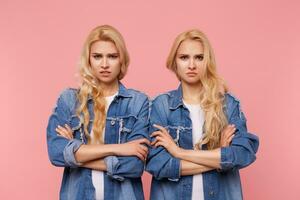 Bewildered young lovely long haired fair-haired sisters keeping hands crossed while looking severely at camera and frowning eyebrows, posing over pink background photo