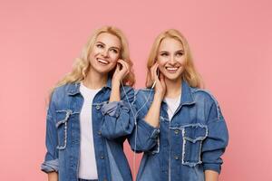 Cheerful young beautiful blonde ladies with wavy hairstyles smiling happily while enjoying music in heaphones while standing over pink background photo