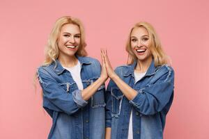 Happy young attractive long haired blonde sisters clapping raised palms of each other and looking with broad smile at camera, standing over pink background photo