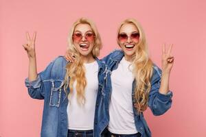 Happy young lovely long haired blonde females in casual clothes forming victory signs with raised fingers while looking excitedly at camera, isolated over pink background photo