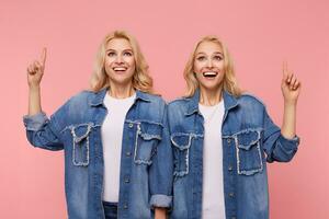 Agitated young lovely blonde sisters dressed in jeans coats and white t-shirts showing excitedly upwards with raised index fingers, standing over pink background photo