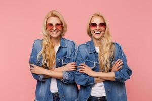 Joyful young lovely long haired blonde twins keeping hands crossed while looking happily at camera with broad smiles, isolated against pink background photo