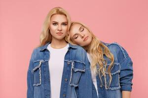 Horizontal photo of young beautiful long haired blonde sisters with curls wearing jeans coats and white t-shirts while standing over pink background with hands down