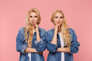 Puzzled young attractive twins curly hairstyle touching face with raised hands and twisting pensively mouth while standing over pink background in jeans coats photo