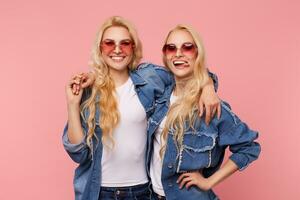 Joyful young pretty long haired blonde sisters in red sunglasses embracing and looking happily at camera with broad smiles, isolated over pink background photo