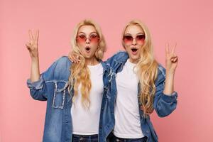 Surprised young attractive white-headed females with wavy hairstyle showing peace gesture and looking amazedly at camera while standing over pink background photo