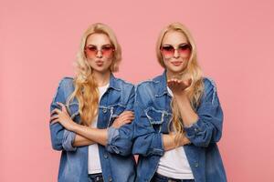 Horizontal photo of young beautiful long haired females with wavy hairstyle folding lips in air kiss while looking positively at camera, isolated over pink background