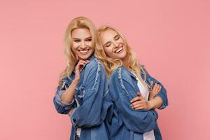 Indoor shot of young pretty fair-haired ladies with wavy hairstyle leaning back to back while posing over pink background in casual wear and laughing cheerfully photo