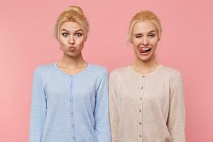 Portrait of beautiful young blonde twins fool around and make faces isolated over pink background. One girl sends a kiss and the second one tongues tooth and winks. photo