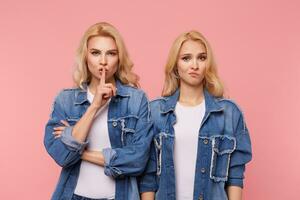 Serious young pretty blonde lady raising hand with hush gesture to her mouth while asking to keep secret, standing over pink background with puzzled fair-haired female photo