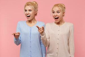 Shocked beautiful young blonde twins with wide open mouth isolated over pink background pointed point to something awesome. photo