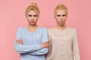 Frowning beautiful young blonde twins in a bad mood, looking in the camera isolated over pink background. photo