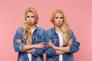 Offended young pretty blonde ladies with curls folding hands on chest while pouting sadly lips, dressed in casual clothes while posing over pink background photo