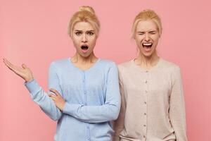 Beautiful young blonde twins are in a bad mood isolated over pink background. One of them is shocked by the news that that upset her sister to tears. photo