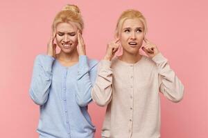 Beautiful young blonde twins heard an unpleasant sound from which there is a ring in the ears and a head splits, isolated over pink background. photo