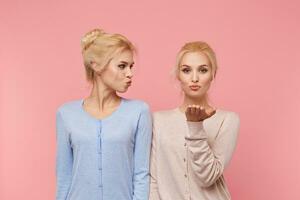 Portrait of attractive blonde young twins sisters, send air kisses, expresses love to someone on distance, stands over pink background. photo