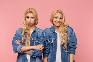 Unhappy young long haired blonde lady looking sadly down while posing over cheerful curly sister with wide smile, isolated against pink background in casual wear photo