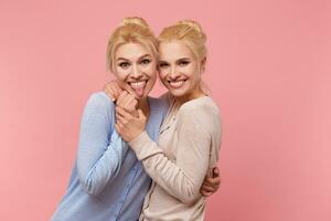 Photos of two sisters, hug each other and hold hands, having fun and smiling widely into the camera, glad that they have each other - they are always fun together Stads over pink background.