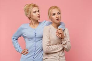 Blonde twin sisters look up in surprise, one of them is clearly dissatisfied with what she sees. Stands over pink background. photo