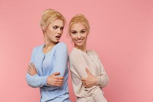 Portrait of cute twins blondes, girl is offended by her sister that she is posing better than her, looks dissatisfied, shes sisters smiles and looks at the camera. Girls stands over pink background. photo