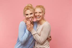 Portrait of cute blonde twins, embraced and held hands, having fun and smiling widely into the camera, stands over pink background. photo