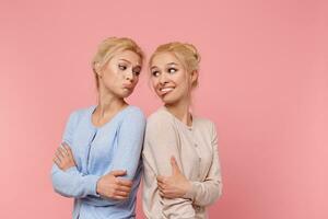 Portrait of cute twins blondes, girls fool standing with their backs to each other, grimace and and look at each other. Stands over pink background. photo