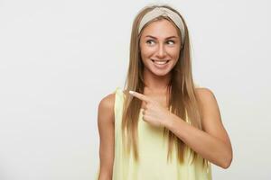 Pleasant looking young joyous long haired blonde lady pointing aside with index finger and smiling happily while standing against white background in beige headband and yellow top photo