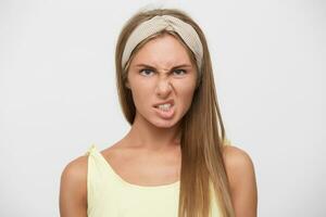 Close-up of displeased young pretty blonde lady with casual hairstyle twisting her mouth and frowning eyebrows while posing over white background, wearing beige headband and yellow top photo