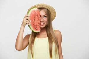 Open-eyed young cheerful pretty blonde lady with casual hairstyle keeping slice of watermelon in raised hand and looking positively at camera, standing over white background photo