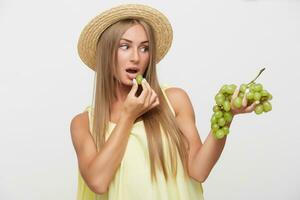 Portrait of beautiful young long haired blonde woman with natural makeup holding bunch of grapes in raised hand and looking excitedly on it, posing over white background photo